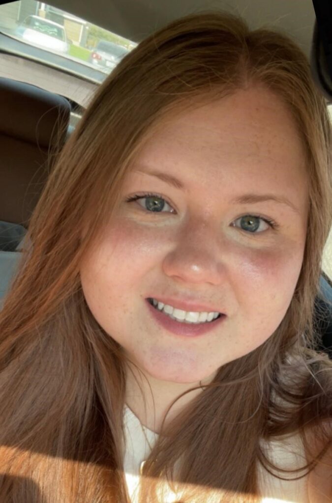 A woman with long light brown hair, possibly one of the sales associates, is sitting in a car, smiling at the camera with sunlight on her face.