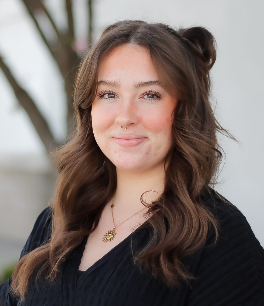 A young woman with long brown wavy hair, a nose ring, and a black top stands outdoors, smiling softly at the camera—her approachable look perfect for sales associates connecting with new customers.