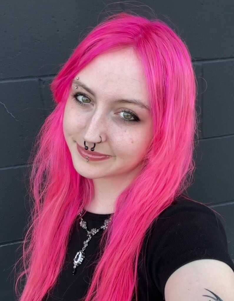 Person with long bright pink hair, facial piercings, and a black shirt standing in front of a dark gray brick wall, looking at the camera and smiling slightly—showcasing the unique style often seen among creative sales associates.