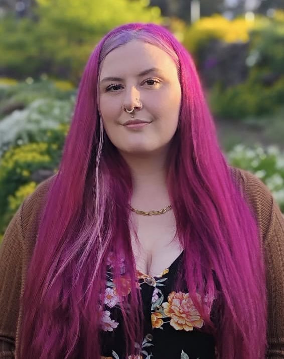 A person with long, bright pink hair, often sought after by body piercers for inspiration, stands outdoors in a garden setting wearing a brown cardigan and floral top.