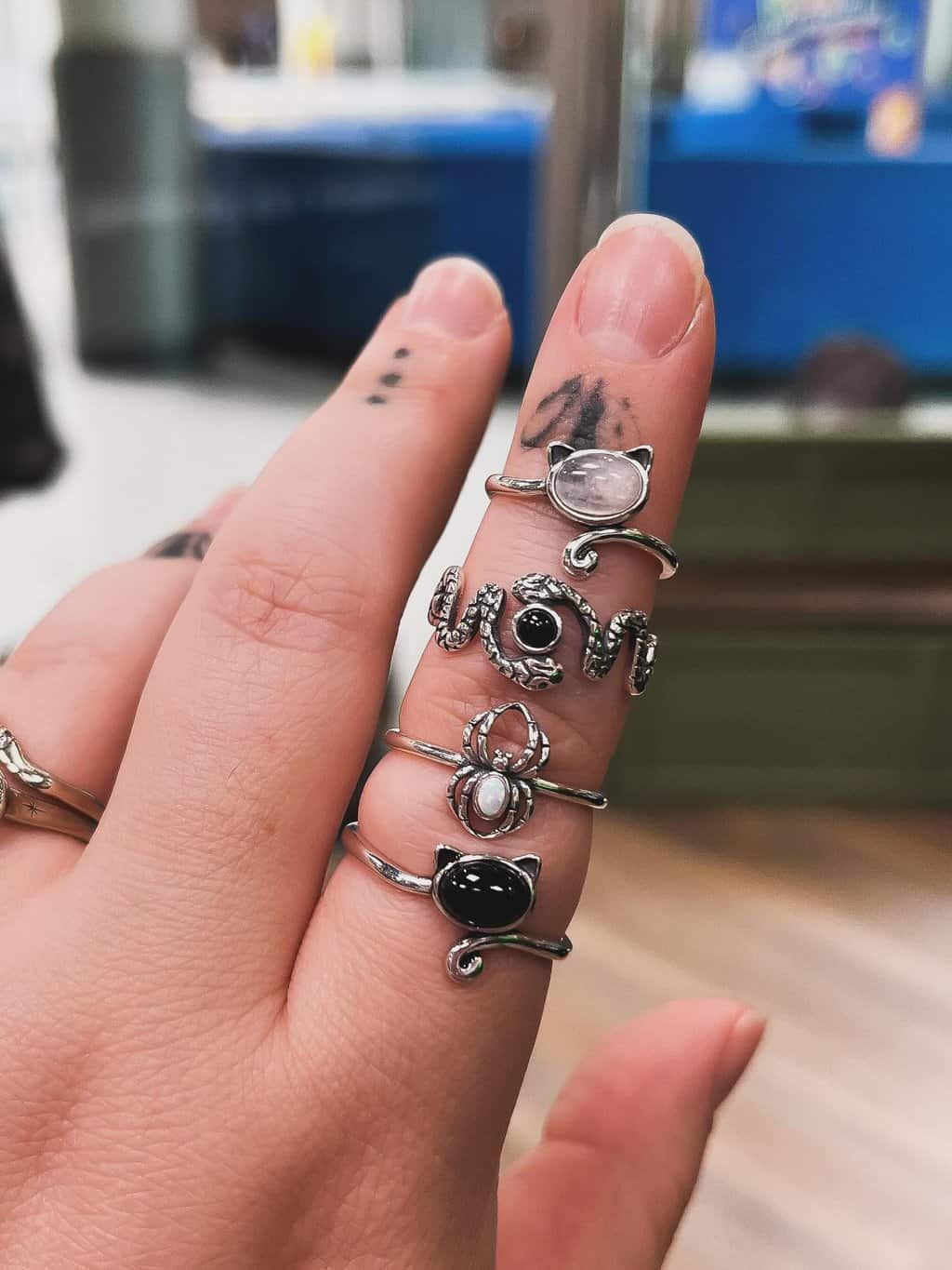 A hand with small tattoos on the fingers wearing four silver rings—featuring unique black and white stones and cat designs on the middle finger—from our women-owned jewelry collection.