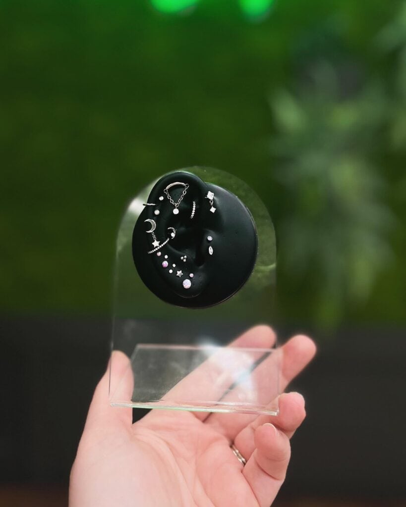 A hand holds a clear stand displaying various small silver earrings and studs on a black circular holder, set against a green blurred background—perfect for showcasing jewelry while discussing store policies with customers.
