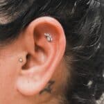 Close-up of a person's ear with two piercings: a small stud on the lobe and a leaf-shaped jewelry piece on the upper cartilage. Dark hair and part of a tattoo are visible.