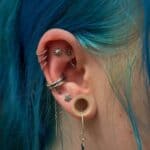 Close-up of an ear with multiple piercings, including rings, studs, a tunnel plug, and dangling earrings; person has vibrant blue hair.