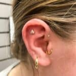 Close-up of an ear with three piercings: a gold hoop in the lobe, a small stud in the helix, and a decorative flower-shaped stud in the inner cartilage.