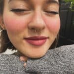 A person with closed eyes and a septum piercing smiles gently, resting their chin on their gray sweater-covered arms.