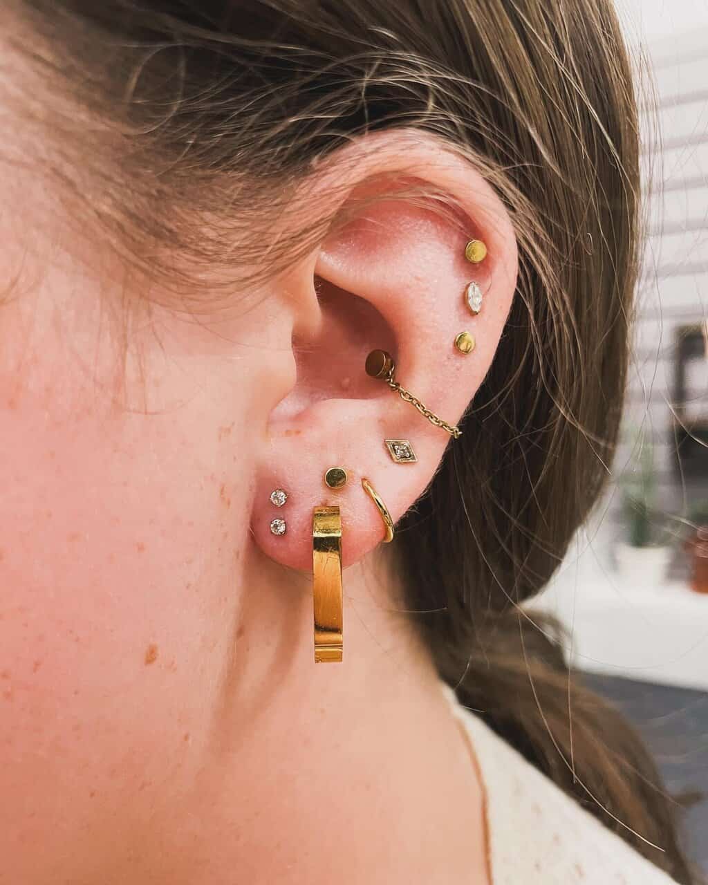 Close-up of an ear with multiple piercings, showcasing gold and diamond stud earrings, hoops, and a chain earring—perfect inspiration and resources for your next curated ear look.