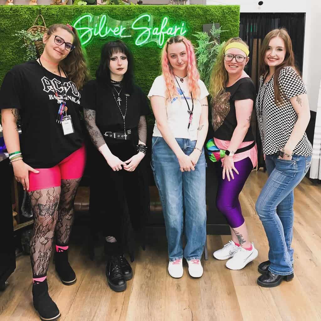 Five women pose for photos indoors in front of a green “Silver Safari” sign, wearing colorful and casual outfits as they stand together on a wooden floor.