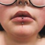 Close-up of a person's lower face with glasses, a septum piercing, and a vertical labret lip piercing.
