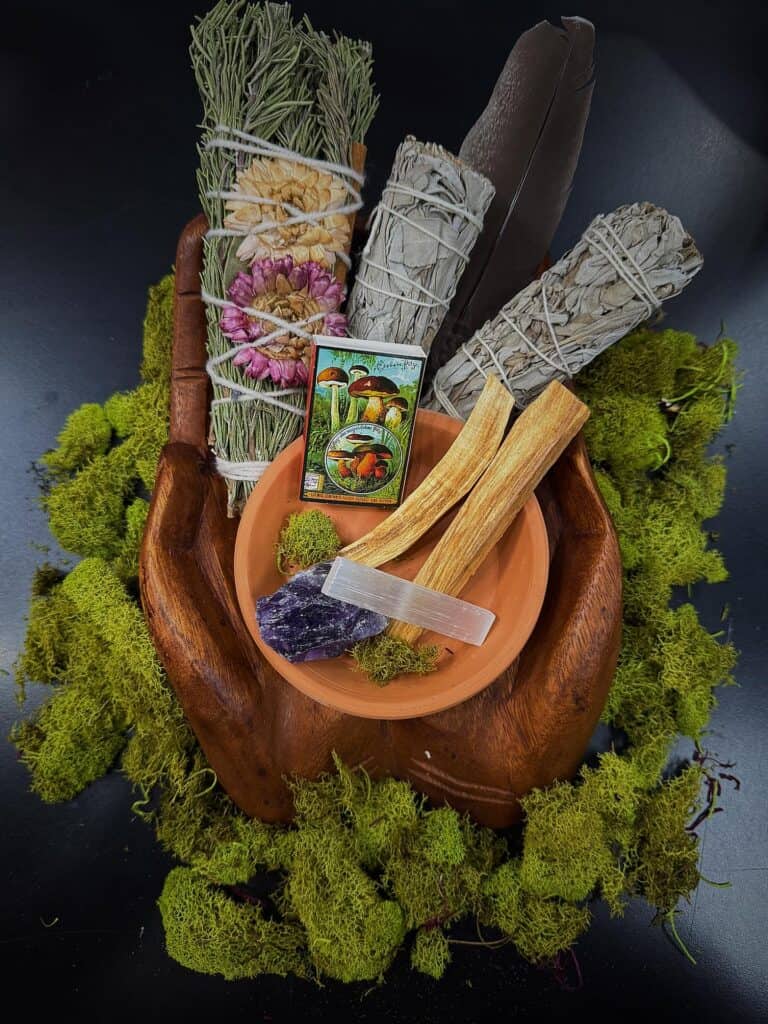 A wooden hand holds sage bundles, a feather, palo santo sticks, crystals, a tarot card, and a clay bowl—all arranged on green moss—inviting reflection on personal rituals and spiritual policies.