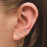 Close-up of a person's ear with three piercings: a bar earring with a chain, and two small stud earrings above it.