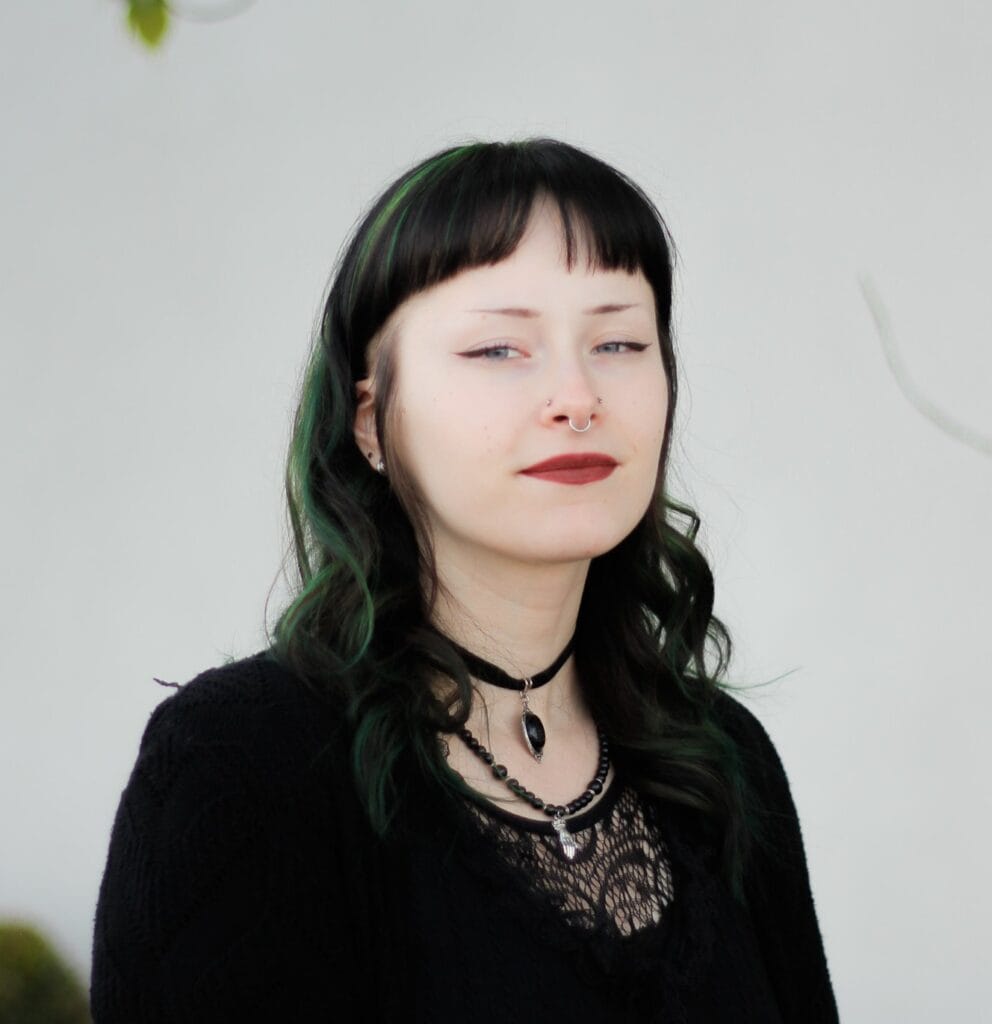 A young woman with pale skin, dark wavy hair with green highlights, wearing black clothing, a choker necklace, and a septum piercing—showcasing the work of skilled body piercers—poses in front of a plain light background.
