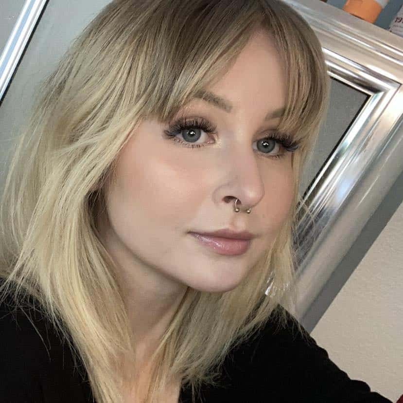 A woman with blonde hair and a septum piercing, dressed casually like many sales associates, poses for a selfie in front of a mirror.