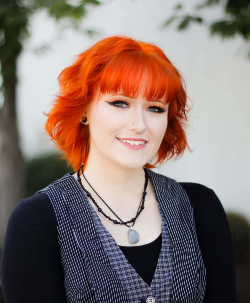 A body piercer with bright orange hair, wearing a black shirt and a pinstriped vest, smiles at the camera.