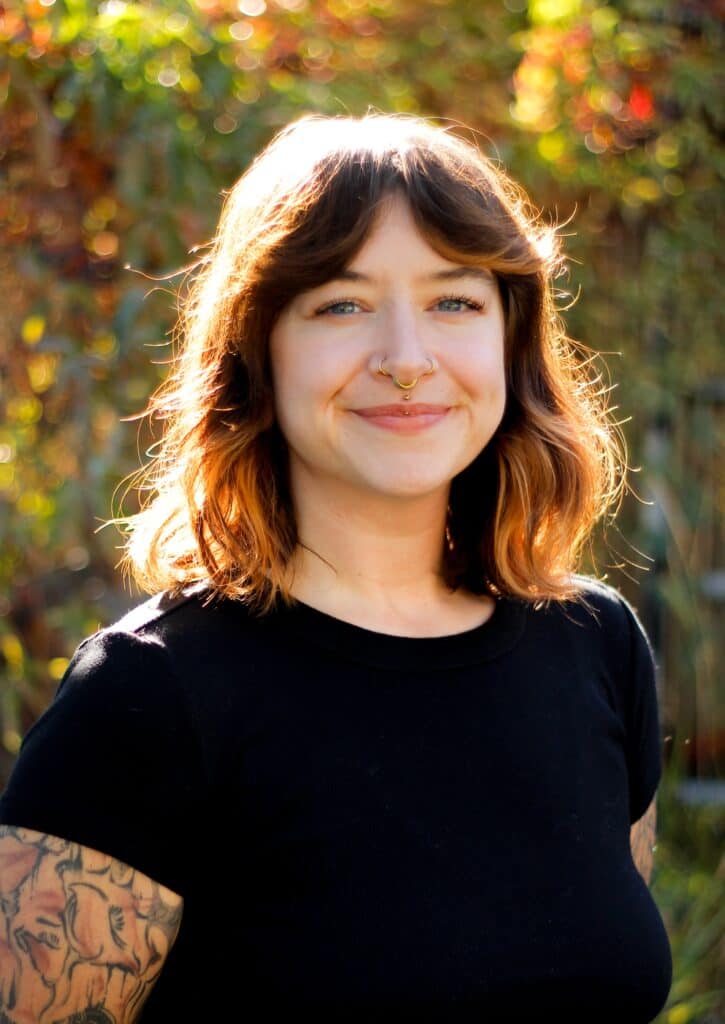 A smiling body piercer with shoulder-length hair, a septum piercing, and arm tattoos stands outdoors.