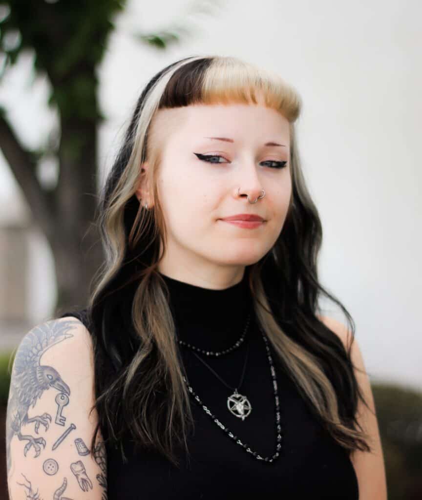 Portrait of a body piercer with a two-toned blonde and black hairstyle, nose rings, and a visible raven tattoo on their arm.